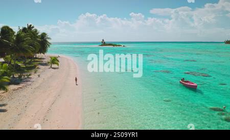 Vista aerea di un turista che cammina lungo una splendida spiaggia di sabbia rosa su un'isola tropicale di aitutaki, isole Cook, con una piccola barca ancorata in acqua turchese Foto Stock