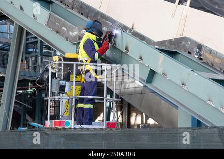 Il saldatore che indossa le attrezzature di sicurezza e utilizza la saldatrice sulla piattaforma aerea salda la struttura metallica Foto Stock