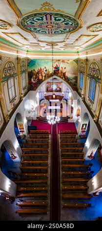 L'immagine mostra l'interno di una chiesa con un soffitto ornato, un grande murale, pali di legno e vetrate colorate. Un lampadario illumina lo spazio. Foto Stock