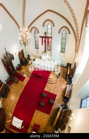 L'immagine cattura una vista aerea dell'interno di una chiesa, evidenziando un altare prominente con un crocifisso, tre vetrate colorate e un tappeto rosso Foto Stock