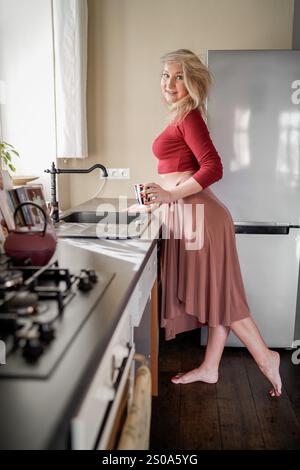 Una donna rilassata e individuale che sta a piedi nudi in una cucina calda mentre tiene in mano una tazza di caffè. Donna che si gusta un caffè mattutino in un gattino luminoso e accogliente Foto Stock
