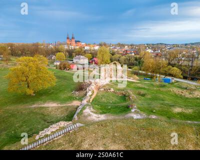 Antiche rovine in un paesaggio erboso con un sentiero, adagiato contro una città caratterizzata da una grande chiesa con due guglie. Sotto sono visibili alberi e piccole case Foto Stock