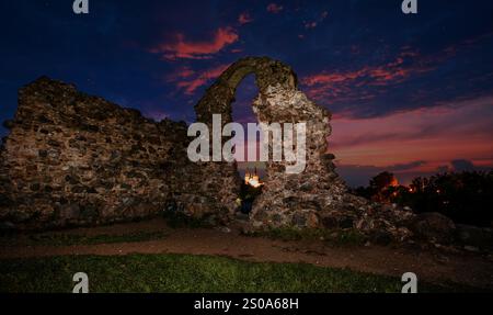 Sagoma di un'antica arcata in pietra contro un vivace cielo serale a Rezekne, Lettonia. Un edificio illuminato con guglie è visibile attraverso l'arco. Foto Stock
