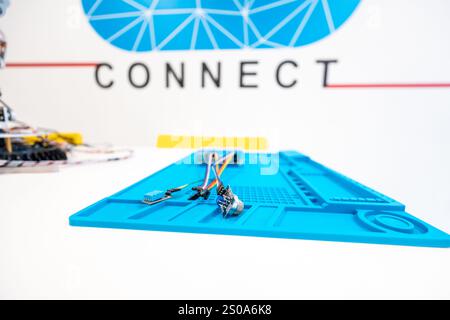 Un tappetino di lavoro blu per componenti elettronici contiene piccoli componenti e fili. Un design geometrico blu e la parola CONNECT sono visibili sulla parete di sfondo. Foto Stock