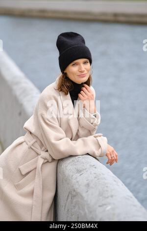Una donna carina si appoggia in modo informale contro una ringhiera accanto a un tranquillo canale navigabile, vestita con un elegante berretto nero e un morbido cappotto beige, godendosi la tranquillità di una Foto Stock
