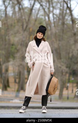 Una donna vestita con un cappotto beige e un berretto nero si posa con sicurezza in un tranquillo ambiente di parco. Gli alberi mostrano segni di autunno, creando un caldo e invitante Foto Stock
