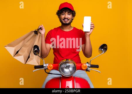Corriere in abbigliamento rosso con borse e telefono, in posa su uno scooter con sfondo giallo brillante Foto Stock