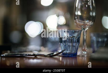 Un primo piano di un bicchiere blu e un bicchiere di vino su un tavolo da pranzo, con luci bokeh soffuse sullo sfondo, a creare un'atmosfera calda e invitante. Foto Stock
