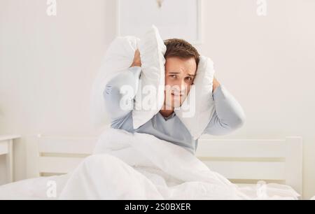 Un uomo senza sonno turbato che copre la testa con cuscini che soffrono di un forte rumore in camera da letto. Foto Stock