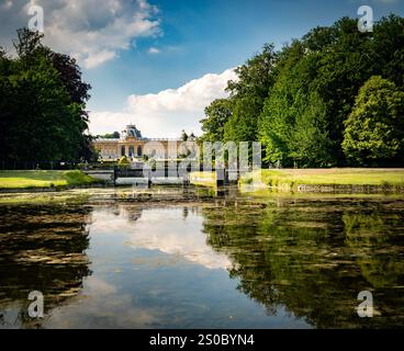 Una vista tranquilla di un parco con un laghetto calmo che riflette il cielo e gli alberi. Sullo sfondo, e' visibile un grandioso edificio, circondato da lussureggiante vegetazione e. Foto Stock