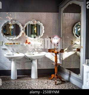 Un bagno elegante con due eleganti lavandini con specchi decorativi, un piedistallo in legno vintage con specchio rotondo e un grande specchio riflettente ornato Foto Stock