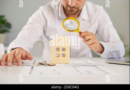 Un giovane uomo d'affari che guarda attraverso una lente d'ingrandimento una casa modello seduta a tavola Foto Stock