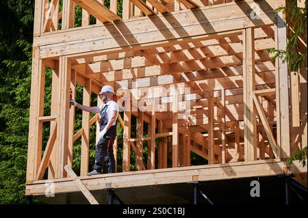 Falegname in procinto di erigere una casa a due piani con struttura in legno vicino ai boschi. L'uomo verifica la planarità del muro con l'aiuto di Level. Concetto di costruzione ecologica contemporanea. Foto Stock