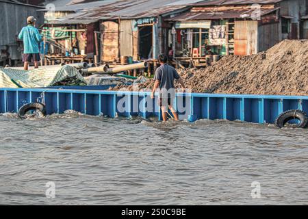 Mekong Delta, Vietnam. 27 dicembre 2024: Un uomo vietnamita cammina in acqua lungo la sua nave da carico/chiatta sovraccarica di sabbia. L'estrazione di sabbia e ghiaia (miniere o draghe) utilizzata per l'industria edile solleva avvertimenti circa l'impatto ambientale dell'eccessivo sfruttamento dei sedimenti dei fiumi (biodiversità e ecosistema), ma anche sulla vita delle persone che vivono sulle rive del fiume. Lo sfruttamento legale o illegale della sabbia provoca una massiccia erosione delle banche, con conseguente perdita di terreni agricoli, crolli di edifici o case e delocalizzazioni della popolazione. Crediti: KEVIN IZORCE/Alamy Live News Foto Stock