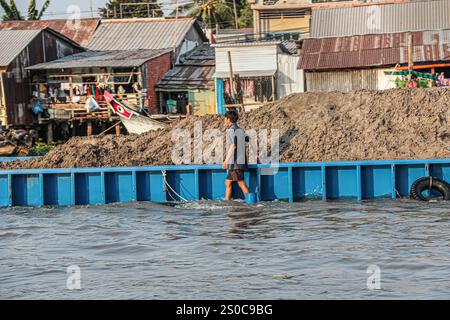 Mekong Delta, Vietnam. 27 dicembre 2024: Un uomo vietnamita cammina in acqua lungo la sua nave da carico/chiatta sovraccarica di sabbia. L'estrazione di sabbia e ghiaia (miniere o draghe) utilizzata per l'industria edile solleva avvertimenti circa l'impatto ambientale dell'eccessivo sfruttamento dei sedimenti dei fiumi (biodiversità e ecosistema), ma anche sulla vita delle persone che vivono sulle rive del fiume. Lo sfruttamento legale o illegale della sabbia provoca una massiccia erosione delle banche, con conseguente perdita di terreni agricoli, crolli di edifici o case e delocalizzazioni della popolazione. Crediti: KEVIN IZORCE/Alamy Live News Foto Stock