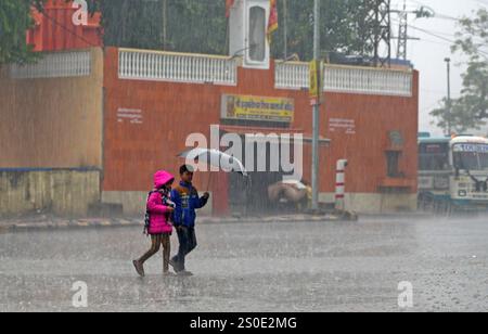 Beawar, Rajasthan, India, 27 dicembre 2024: Bambini che tengono un ombrello a piedi su strada in mezzo alla pioggia la mattina d'inverno a Beawar. La vita quotidiana nel Rajasthan è stata gravemente colpita dalla pioggia e dalla nebbia che hanno colpito lo stato, portando a un brusco calo delle temperature e a una ridotta visibilità in diverse aree. Il tempo inclemente ha intensificato il freddo, con una fitta nebbia che ricopre molte regioni. L'India Meteorological Department (IMD) ha emesso un allarme arancione per 15 distretti e un allarme giallo per sei distretti nello stato, avvertendo della persistente nebbia densa nei prossimi tre giorni. Crediti: Sumit Saraswat / A. Foto Stock