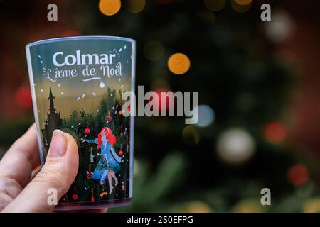 Colmar, Francia - 14 dicembre 2024: Coppa di VIN brulé in mano donna, Colmar L ame de Noel su di esso, luci del mercatino di Natale e un albero sullo sfondo Foto Stock