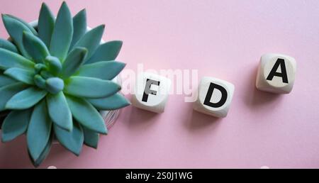 FDA - simbolo della Food Drug Administration. Cubi di legno con la parola FDA. Bellissimo sfondo rosa con piante succulente. Amministrazione di farmaci medicali e alimentari Foto Stock