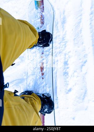 Una foto dal punto di vista di uno snowboarder che indossa pantaloni gialli e stivali neri, in piedi su uno snowboard colorato. Foto Stock