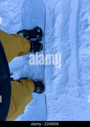 Una foto dal punto di vista di uno snowboarder con pantaloni gialli e stivali neri che cavalcano su una pista innevata. Foto Stock
