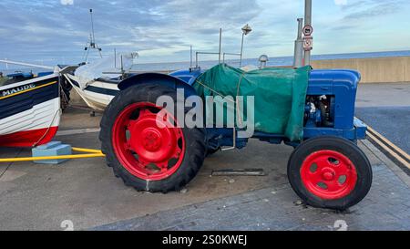 Un trattore blu con ruote rosse, parzialmente coperto da un telone verde, è parcheggiato vicino a una piccola barca da pesca chiamata "Maude". La scena è ambientata all'aperto da l Foto Stock