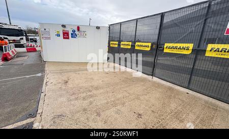 Un'entrata in cantiere, caratterizzata da un edificio bianco temporaneo, una recinzione a rete nera con cartello 'FARRANS' e un'area pavimentata. Un carrello è parzialmente Foto Stock
