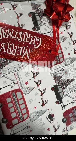 Primo piano di un regalo di Natale confezionato in carta bianca con un tema londinese caratterizzato da dettagli rossi glitter su autobus, renne e un cartellino regalo. Un fiocco rosso a. Foto Stock