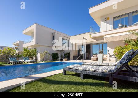 Elegante villa a Vistabella, Spagna, con piscina privata, vegetazione lussureggiante, lettini e architettura moderna sotto un cielo blu. Foto Stock