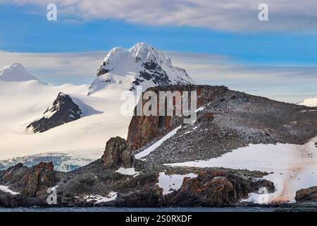 Costa della penisola antartica, mattina presto. Costa rocciosa con pinguini; dietro montagne innevate; nuvole, cielo blu sopra. Foto Stock