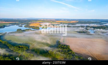 La luce nebulosa del mattino proietta un'atmosfera serena sullo stagno di Rozmberk, vicino a Trebon, in Cechia. La nebbia si libra sopra l'acqua, creando un paesaggio tranquillo con vegetazione lussureggiante e campi lontani. Foto Stock