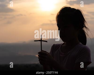 Silhouette di una bambina carina che tiene una croce in preghiera da sola in un parco contro un caldo tramonto. Foto Stock