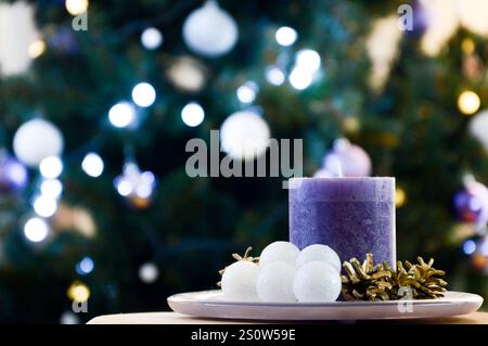 Una candela viola circondata da piccole palline di natale bianche e coni di pino dorato si trova di fronte a un albero di Natale splendidamente illuminato. bokeh, offuscato Foto Stock