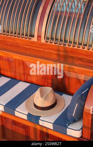 Elegante cappello di paglia poggiato su un cuscino a righe blu e bianche a bordo di un classico yacht a vela. Foto Stock