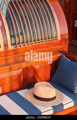 Elegante cappello di paglia poggiato su un cuscino a righe blu e bianche a bordo di un classico yacht a vela. Foto Stock