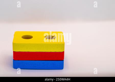 Tre blocchi di legno colorati impilati verticalmente. Il blocco superiore è giallo con due fori, il centro è rosso e il basso è blu. Sono su un whi Foto Stock