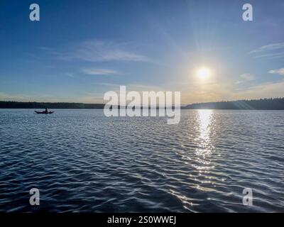 Un kayak solitario pedala su un lago sereno sotto un tramonto morbido, circondato da colline boscose lontane e da un'atmosfera tranquilla. Foto Stock
