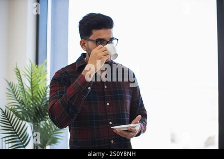 Giovane indiano che indossa occhiali, sorseggiando un caffè alla finestra. Momenti di relax catturati in un ambiente luminoso che trasmette calore e tranquillità. Foto Stock