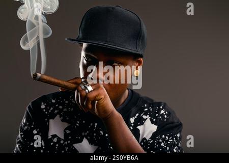 Il giovane con un berretto da baseball e anelli fuma un sigaro e indossa una felpa alla moda su sfondo grigio, che rappresenta la gioventù contemporanea Foto Stock