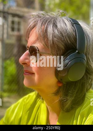 Una donna matura e bella alla moda siede in un giardino primaverile, indossa una moderna giacca verde e occhiali da sole, trasudando sicurezza e stile in mezzo al b Foto Stock
