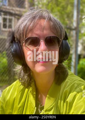 Ritratto di donna matura e alla moda siede in un giardino primaverile, indossando una moderna giacca verde e occhiali da sole, trasudando sicurezza e stile in mezzo al b Foto Stock