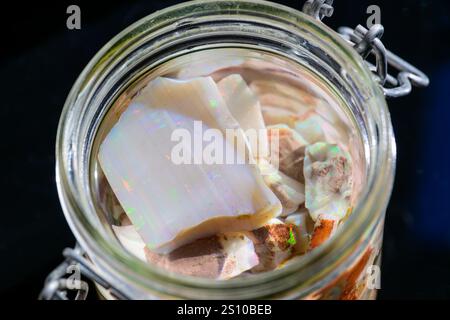 Un vaso di vetro pieno di vari pezzi di opale e frammenti minerali, che mostrano i loro colori e le loro texture uniche. Il vaso è sigillato con un ciondolo metallico Foto Stock