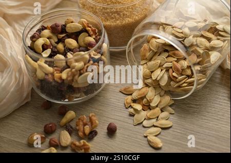 Zucchero di canna da zucchero, mix di noci, uvetta e semi di zucca sparsi in vasetti di vetro trasparenti su tavola di legno, panno beige sullo sfondo Foto Stock