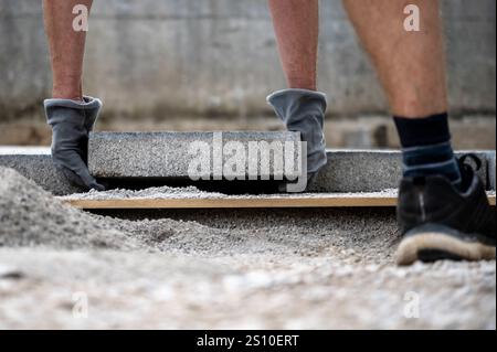 Un operatore che indossa i guanti posiziona con cura una lastra di cemento su una base di ghiaia preparata. Il primo piano cattura la precisione e lo sforzo durante la costruzione Foto Stock