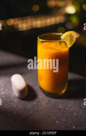 Bicchiere di succo di mango fresco con spicchio di lime su un bancone da bar scuro, illuminazione soffusa, con sfondo bokeh nella notte buia Foto Stock