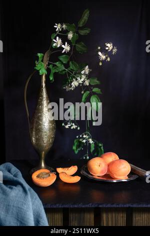 Albicocche fresche in cesto di metallo sul tavolo con fiori in vasi di bronzo su sfondo scuro - fotografie scure e voluminose Foto Stock