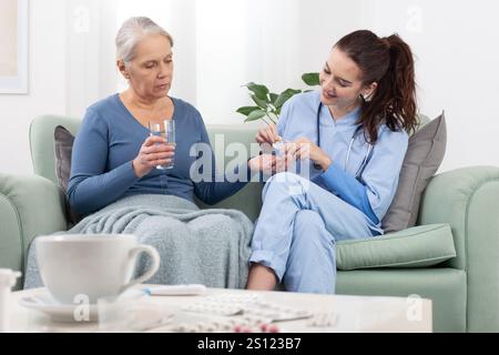 Un infermiere o un medico giovane e amichevole sorride calorosamente mentre aiuta una paziente anziana a prendere una pillola mentre si siede su un divano a casa, in una clinica medica, Foto Stock
