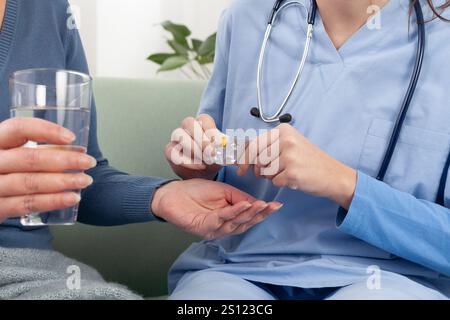 Primo piano delle mani di un infermiere o di un medico che aiuta una paziente anziana a prendere una pillola mentre si siede su un divano a casa, in una clinica medica o a hosp Foto Stock