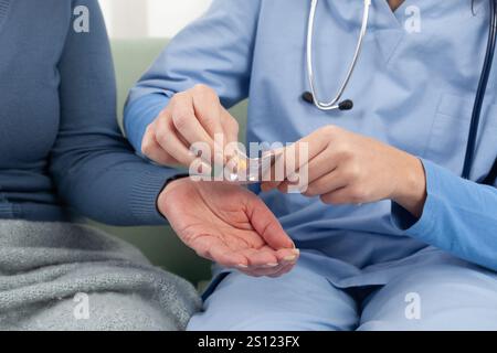 Primo piano delle mani di un infermiere o di un medico che aiuta una paziente anziana a prendere una pillola mentre si siede su un divano a casa, in una clinica medica o a hosp Foto Stock