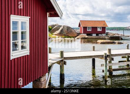 Due boathouses dipinte di rosso e un molo di legno sulla costa di granito rosa nella provincia di Bohuslän, Svezia, sotto un cielo tempestoso in una soleggiata giornata estiva. Foto Stock