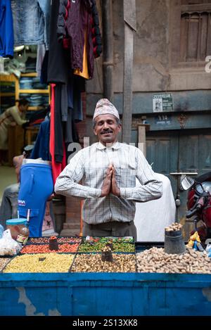Venditore di strada nepalese con tradizionale cappello topi di dacca che esegue un gesto di saluto namaste al suo banco vendendo vari tipi di frutta secca e noci Foto Stock